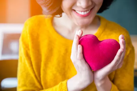 Mulher sorridente com suéter amarelo segurando um coração de pelúcia nas duas mãos