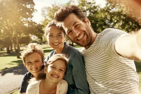 Selfie de uma família de 4 pessoas: o filho e a filha são abraçados pela mãe ao lado do pai, enquanto ele tira a foto.